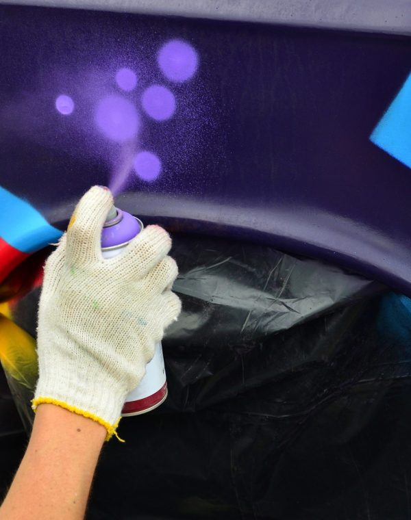 A hand with a spray can that draws a new colorful graffiti on the car. Photo of the process of drawing a graffiti on an automobile close-up. The concept of street art and illegal vandalism
