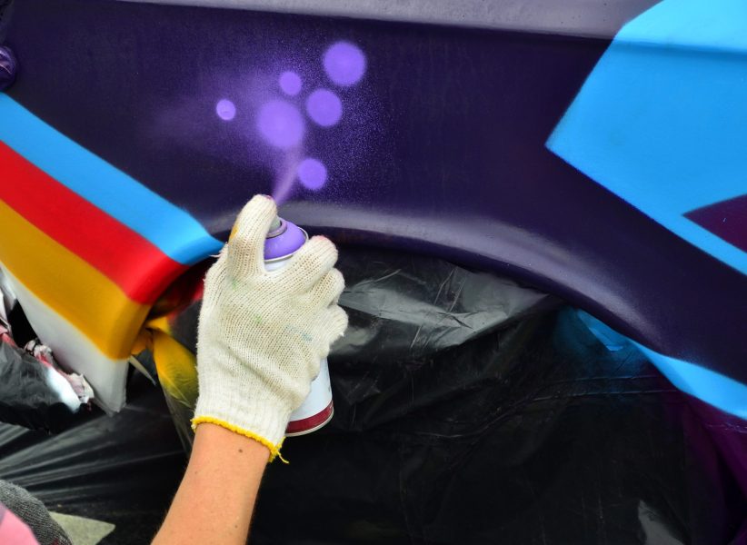 A hand with a spray can that draws a new colorful graffiti on the car. Photo of the process of drawing a graffiti on an automobile close-up. The concept of street art and illegal vandalism